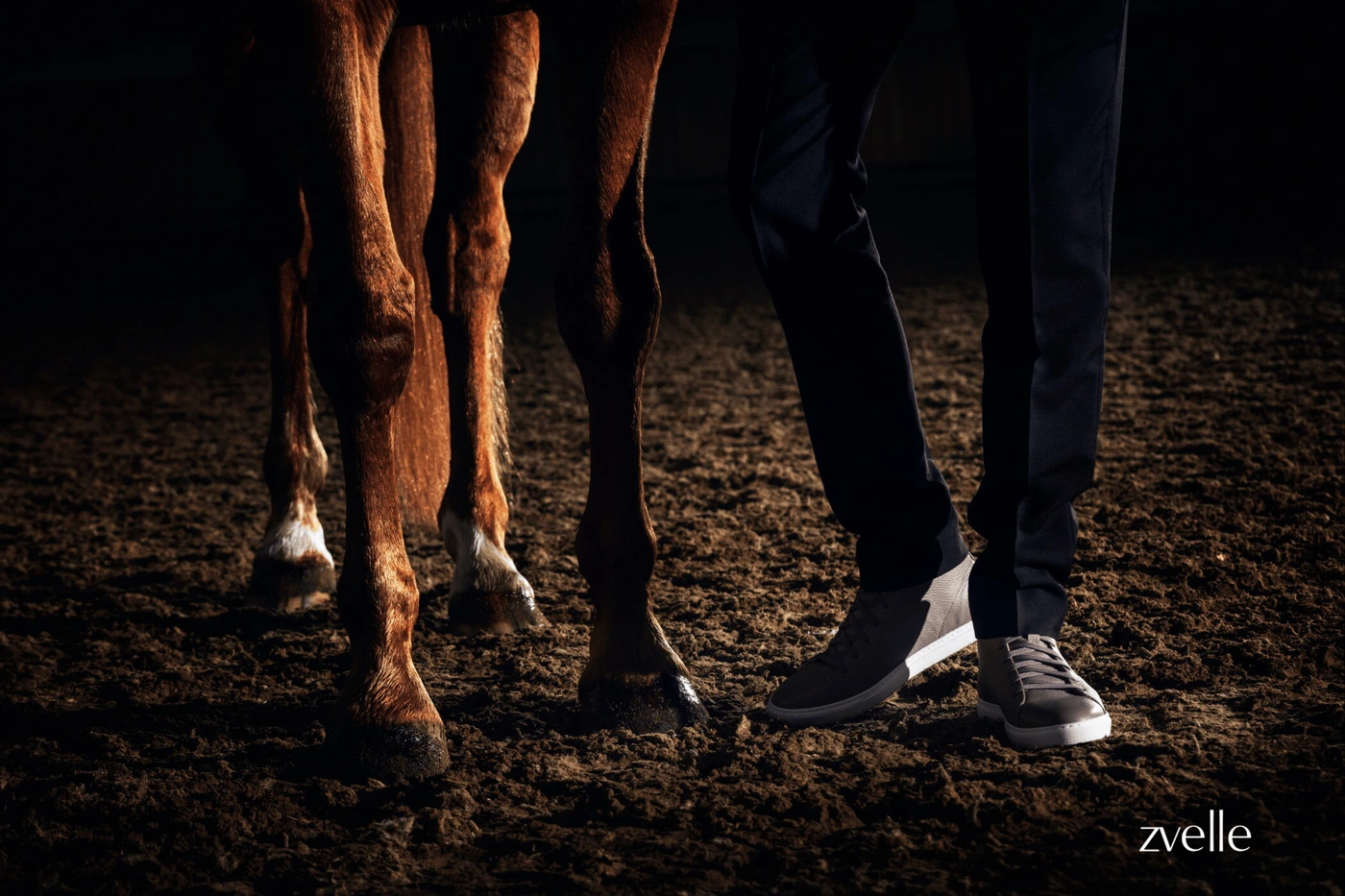 Low-angle shot focusing on a person's legs wearing grey sneakers and dark trousers standing beside a horse on a dirt surface.