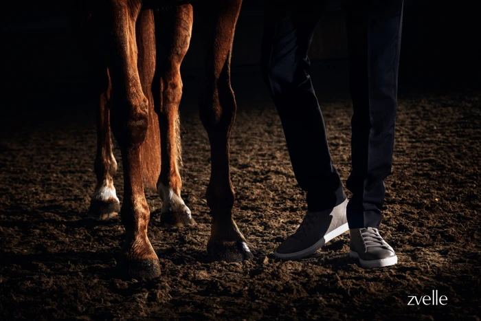 Low-angle shot focusing on a person's legs wearing grey sneakers and dark trousers standing beside a horse on a dirt surface.