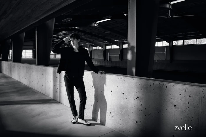 Man wearing dark jacket and trousers leaning against a concrete wall inside an industrial building interior