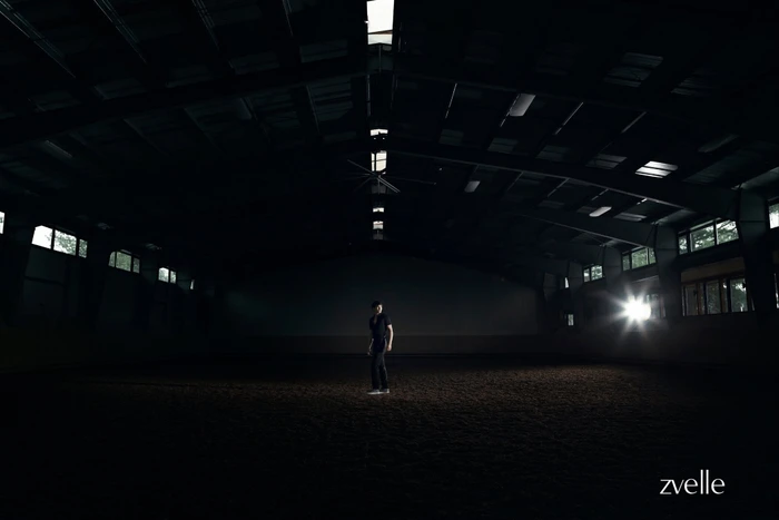 A person standing alone in the center of a large, dark indoor arena lit by a bright light source on the right.