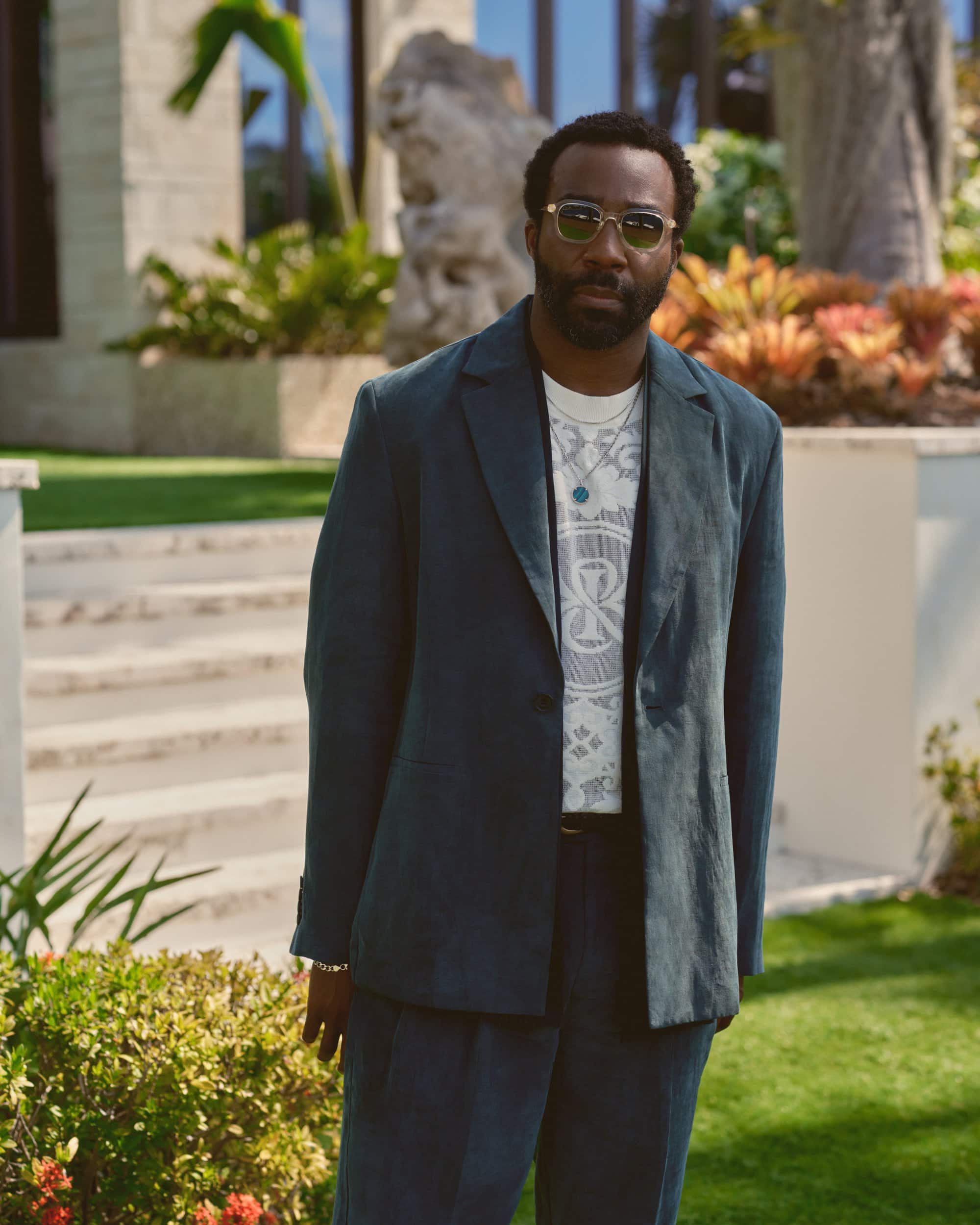 Tramell Tillman wearing a dark blazer over a graphic t-shirt and sunglasses outdoors in an upscale residential setting.