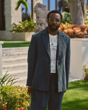 Tramell Tillman wearing a dark blazer over a graphic t-shirt and sunglasses outdoors in an upscale residential setting.