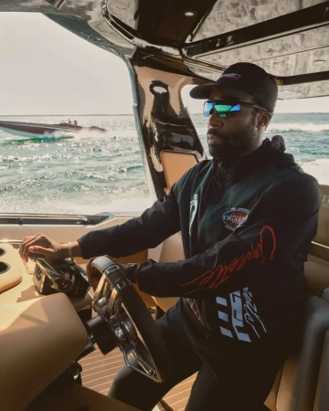 Black man wearing sunglasses and a baseball cap driving a luxury speedboat during the Kith Spring 2025 ad campaign.