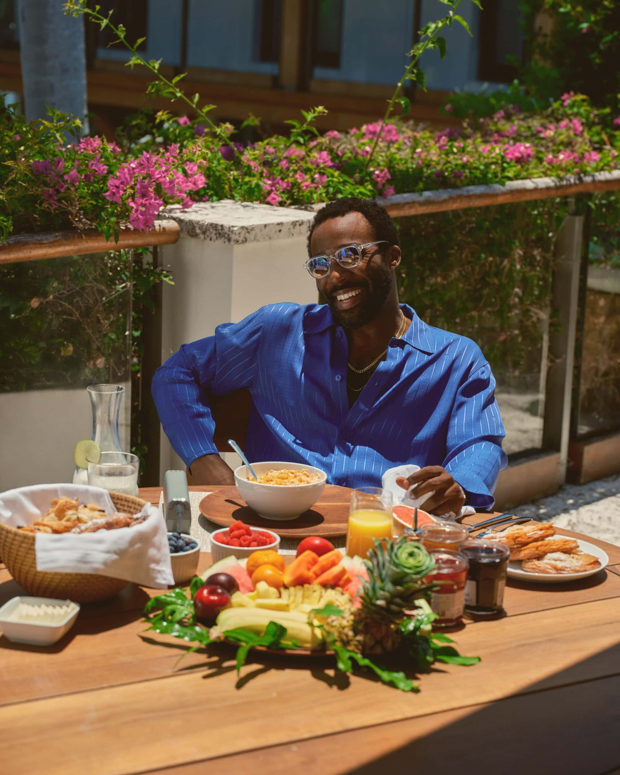 Tramell Tillman wearing a blue pinstripe shirt seated at an outdoor breakfast table filled with fruit and pastries for Kith Spring 2025.