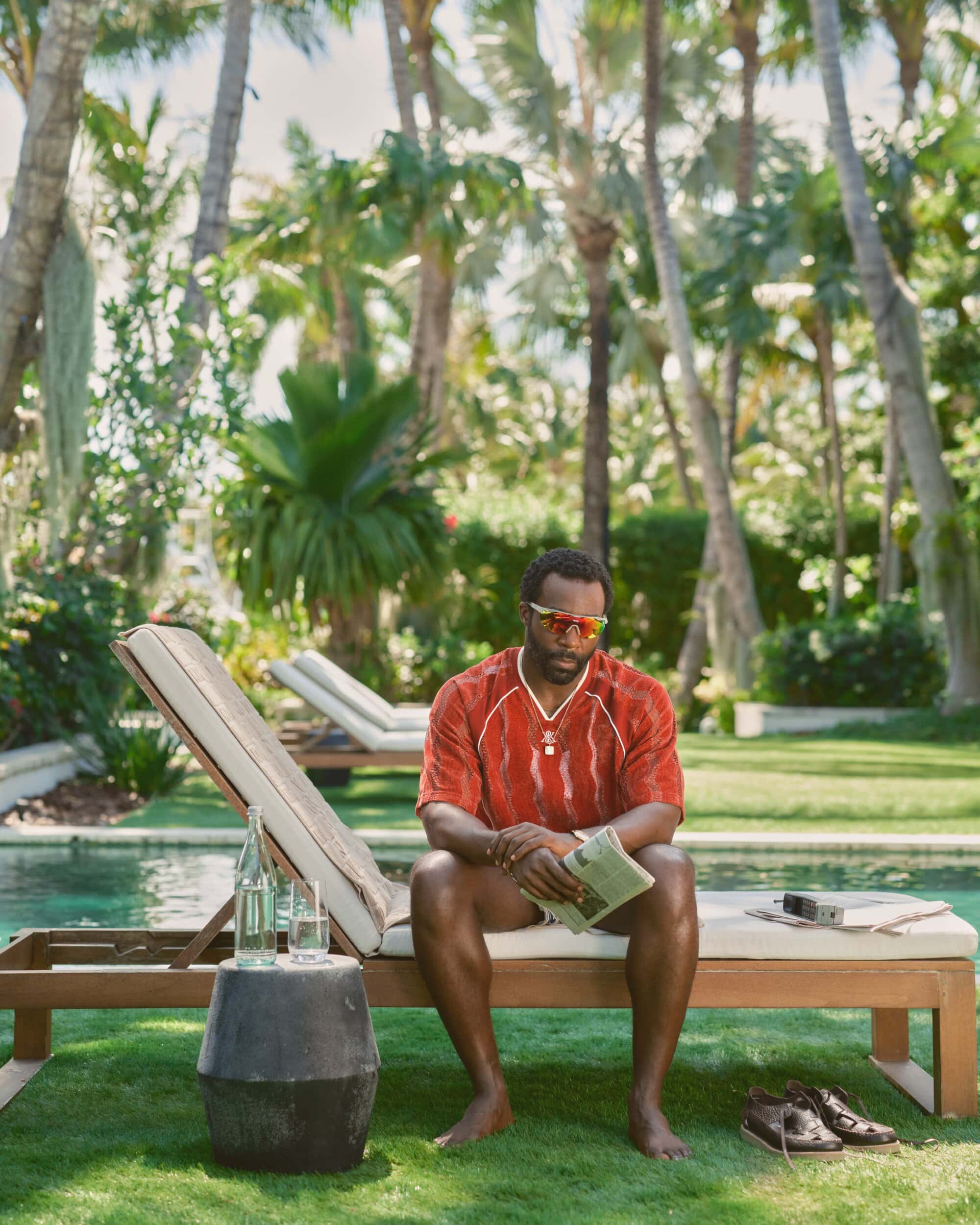 Black man wearing a red athletic jersey and sunglasses reading a newspaper while seated on a lounge chair by a swimming pool.