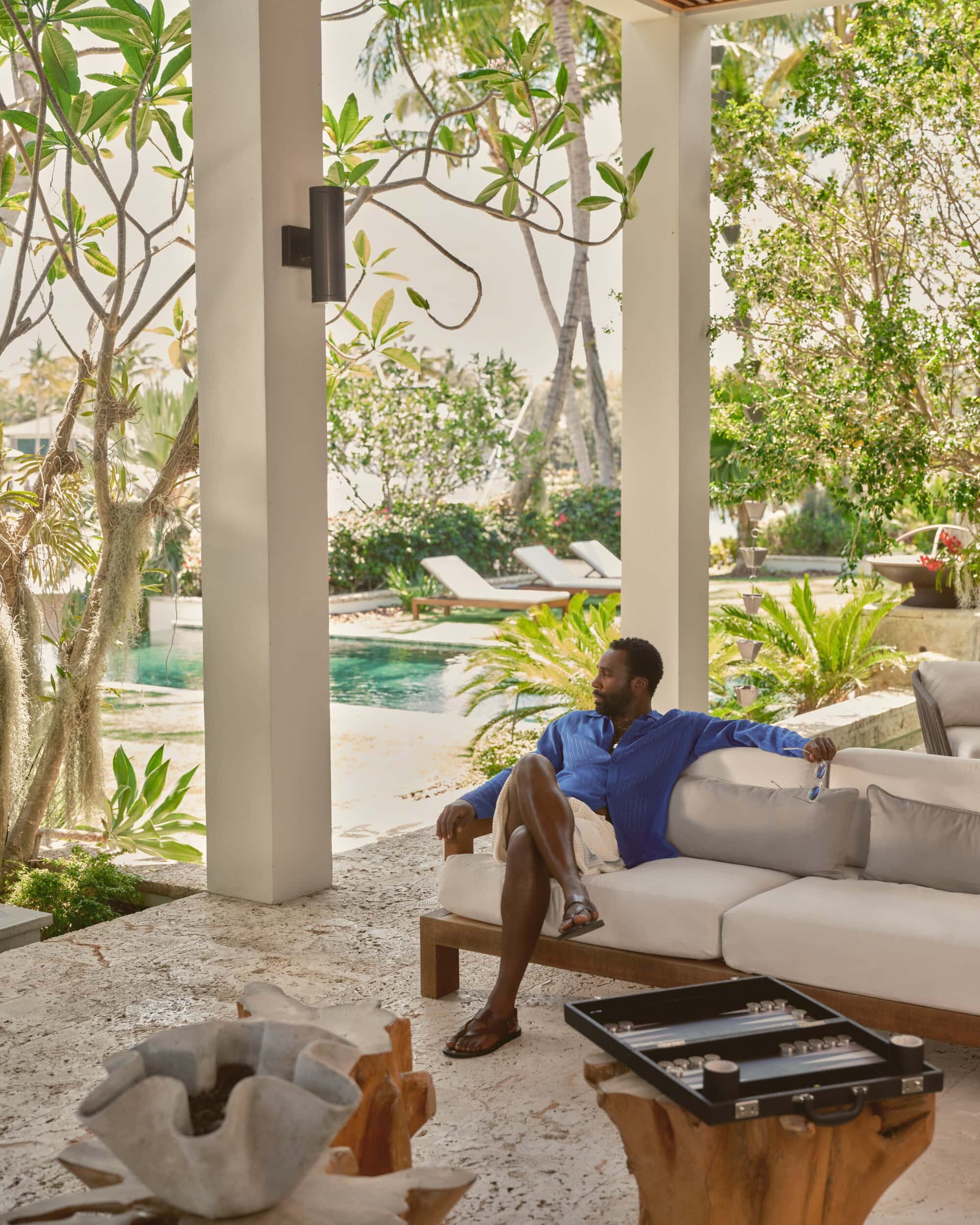 Tramell Tillman seated on an outdoor white sofa wearing a blue shirt and white shorts in a luxury resort patio setting