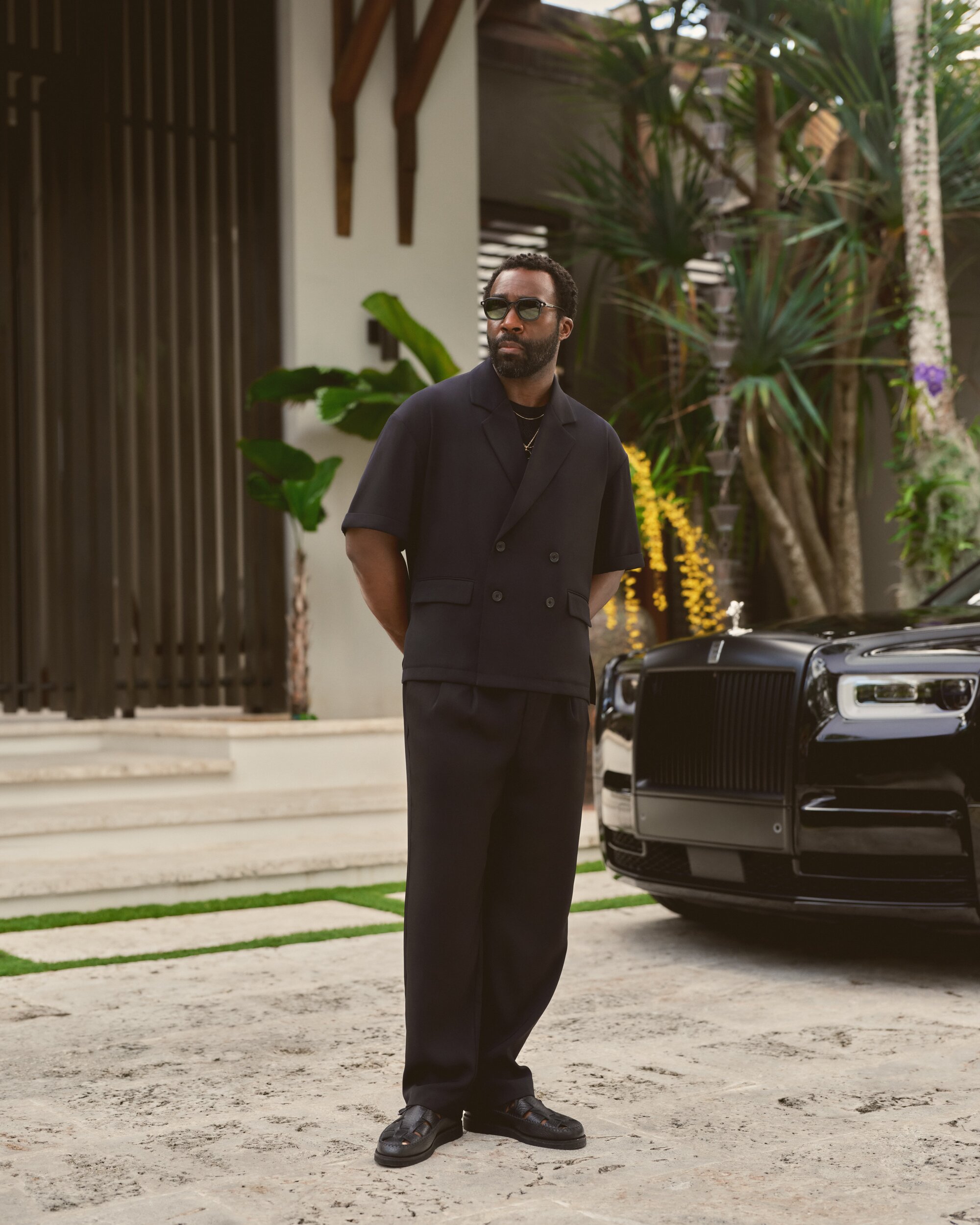 Tramell Tillman wearing a dark short-sleeved blazer and trousers standing beside a black luxury car outdoors.