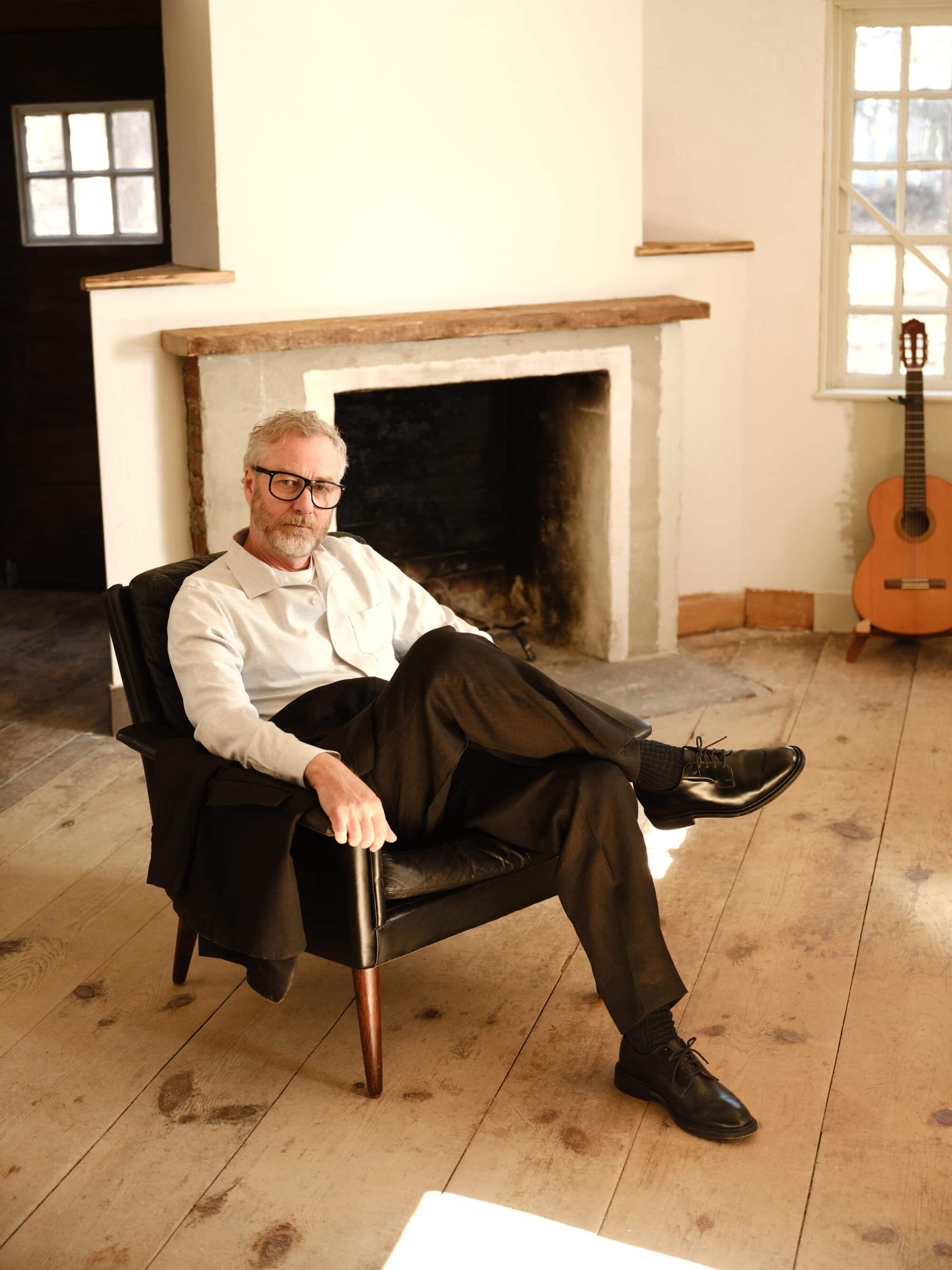 Matt Berninger seated in a black armchair inside a rustic room with a fireplace and an acoustic guitar for the Todd Snyder spring 2025 campaign.