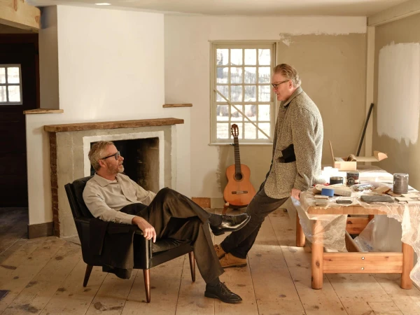 Two men interacting in a rustic room with a fireplace, wooden floor, and a table with paint supplies.