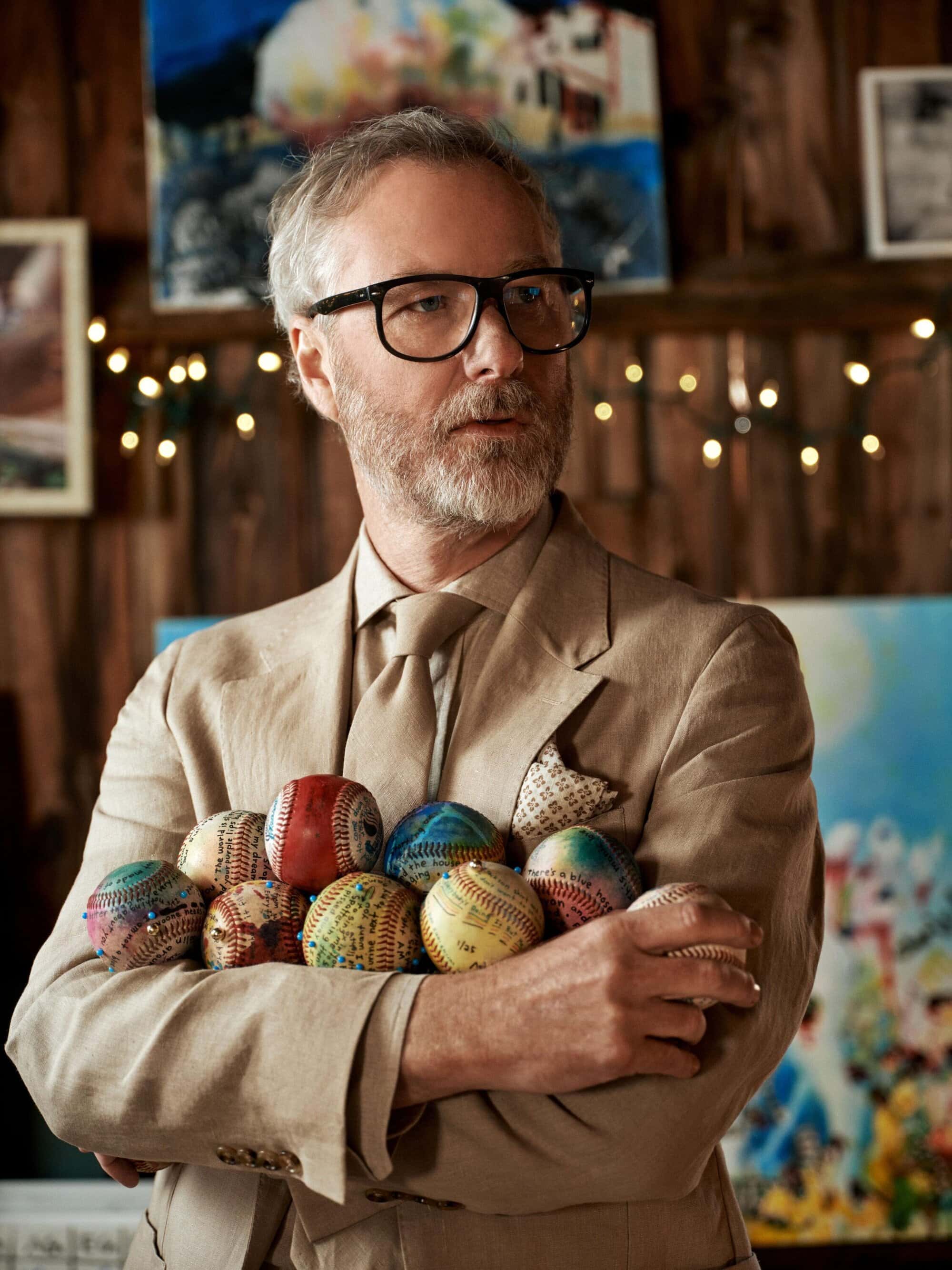 Matt Berninger wearing a tailored beige suit and holding a collection of baseballs in an indoor setting.