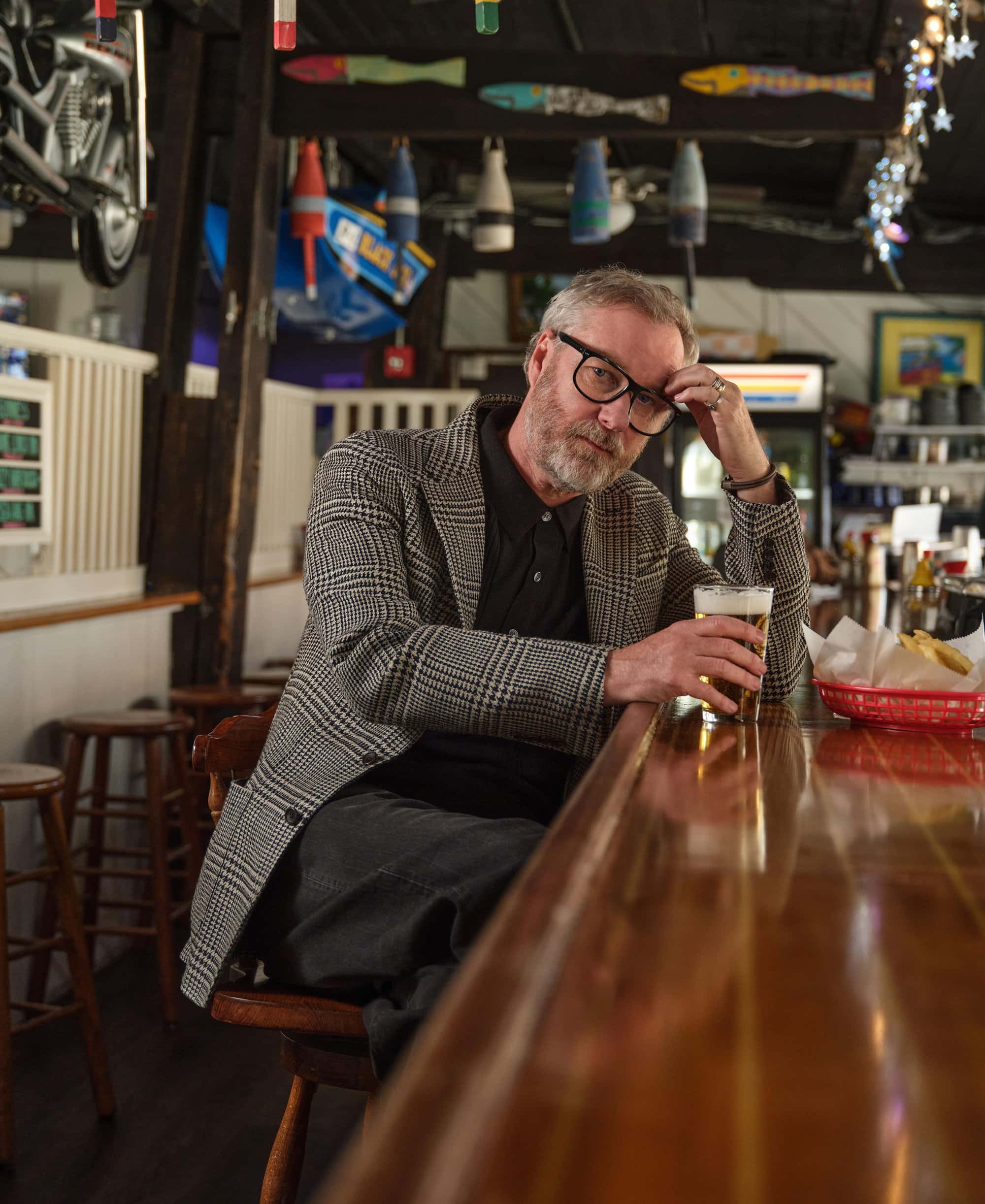 Matt Berninger wearing a plaid blazer and glasses seated at a wooden bar holding a glass of beer for Todd Snyder.