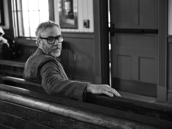 Man wearing glasses and a blazer seated on a wooden bench looking back in a black and white interior setting