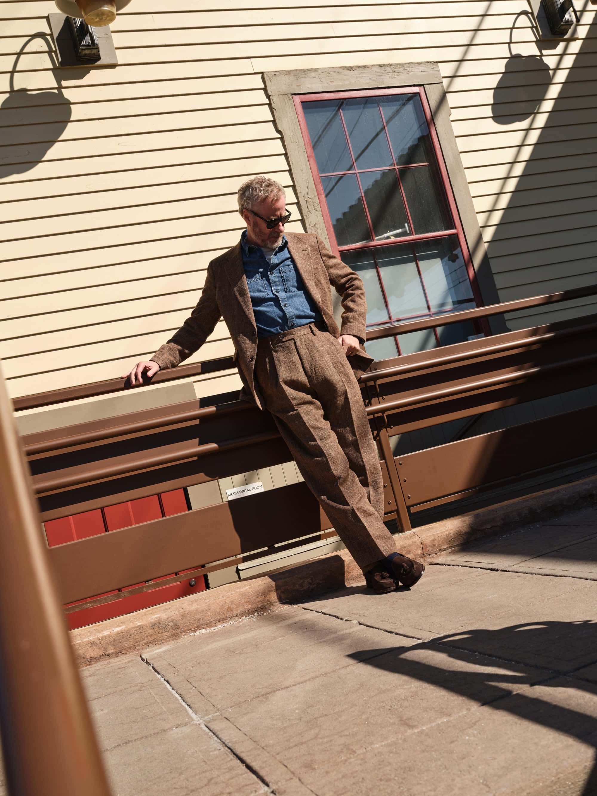 Man wearing a brown suit and sunglasses leaning against a brown railing outside a yellow building.