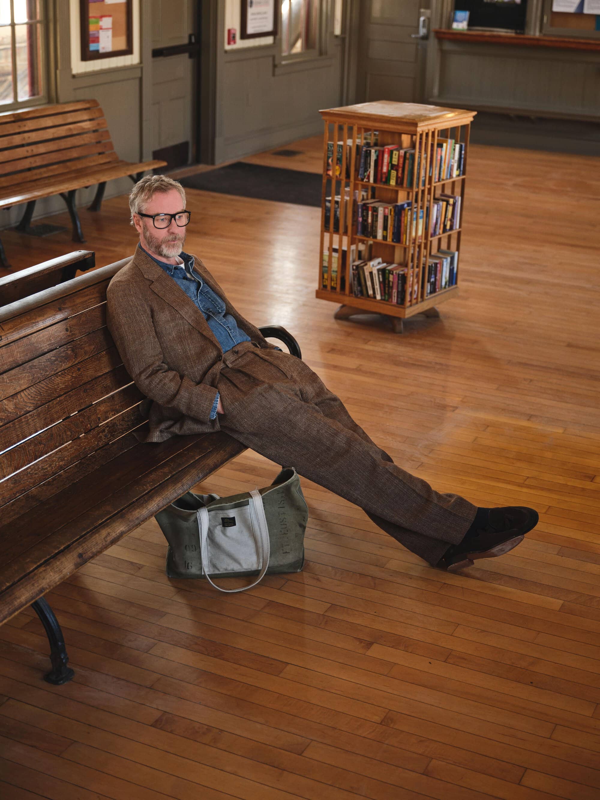 Man wearing a brown tweed suit and glasses relaxing on a wooden bench inside a waiting room with a book rack.