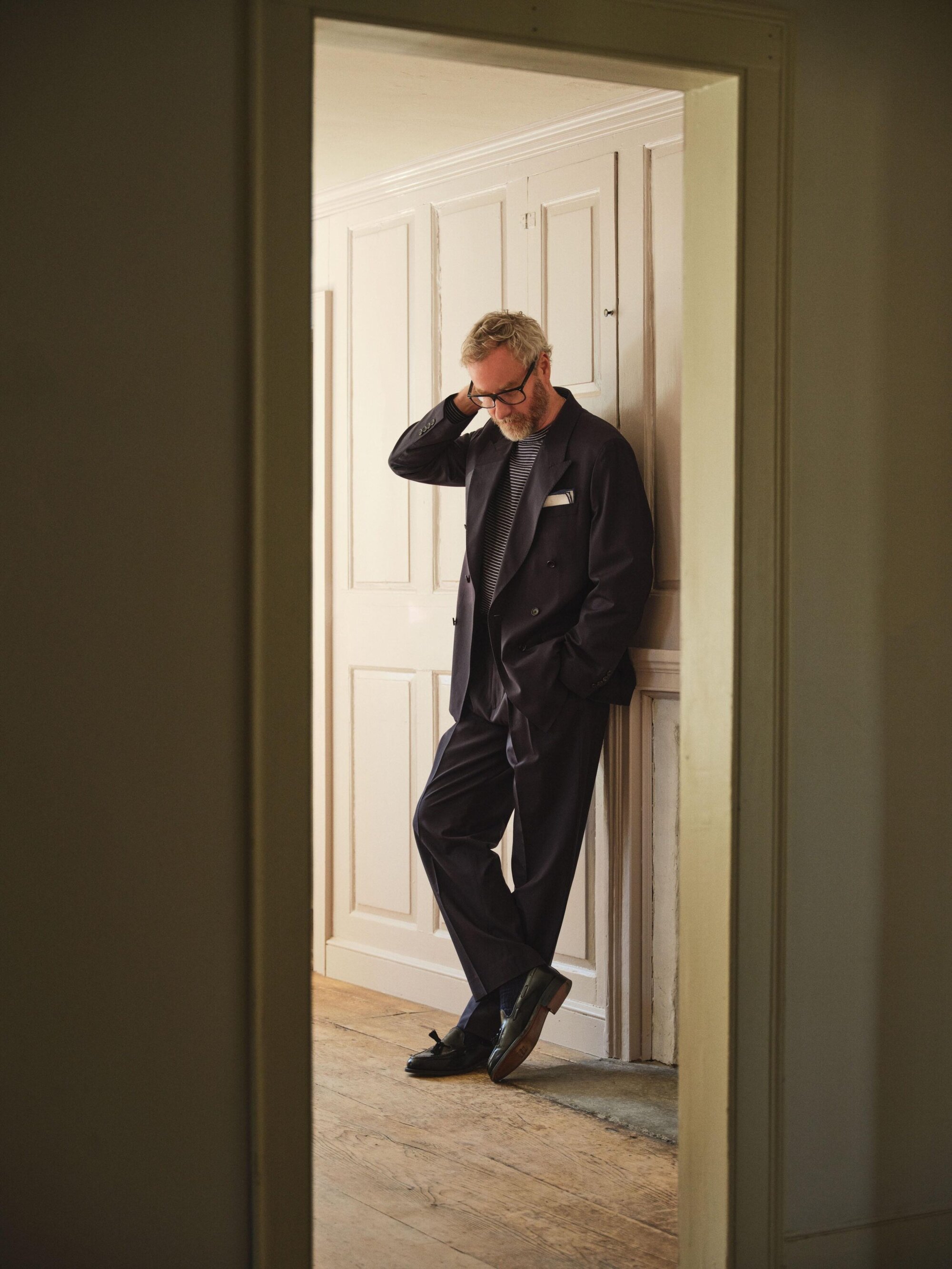 Matt Berninger wearing a dark suit leaning against white panelled walls for Todd Snyder Spring 2025 ad campaign.