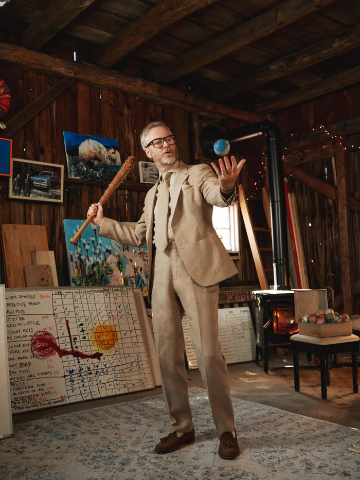 Man wearing a beige suit holding a baseball bat inside a rustic wooden barn with artwork and a wood stove.