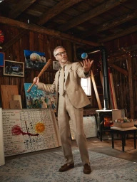 Man wearing a beige suit holding a baseball bat inside a rustic wooden barn with artwork and a wood stove.