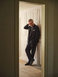 Matt Berninger wearing a dark suit leaning against white panelled walls for Todd Snyder Spring 2025 ad campaign.