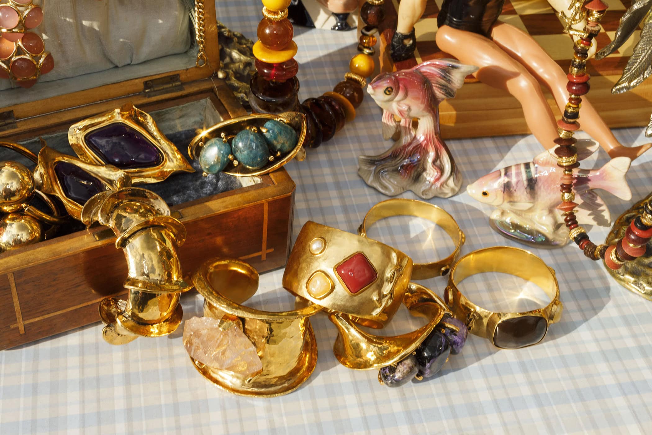 Close-up still life of chunky gold bracelets, gemstone rings, and decorative ceramic fish on a checkered tablecloth.