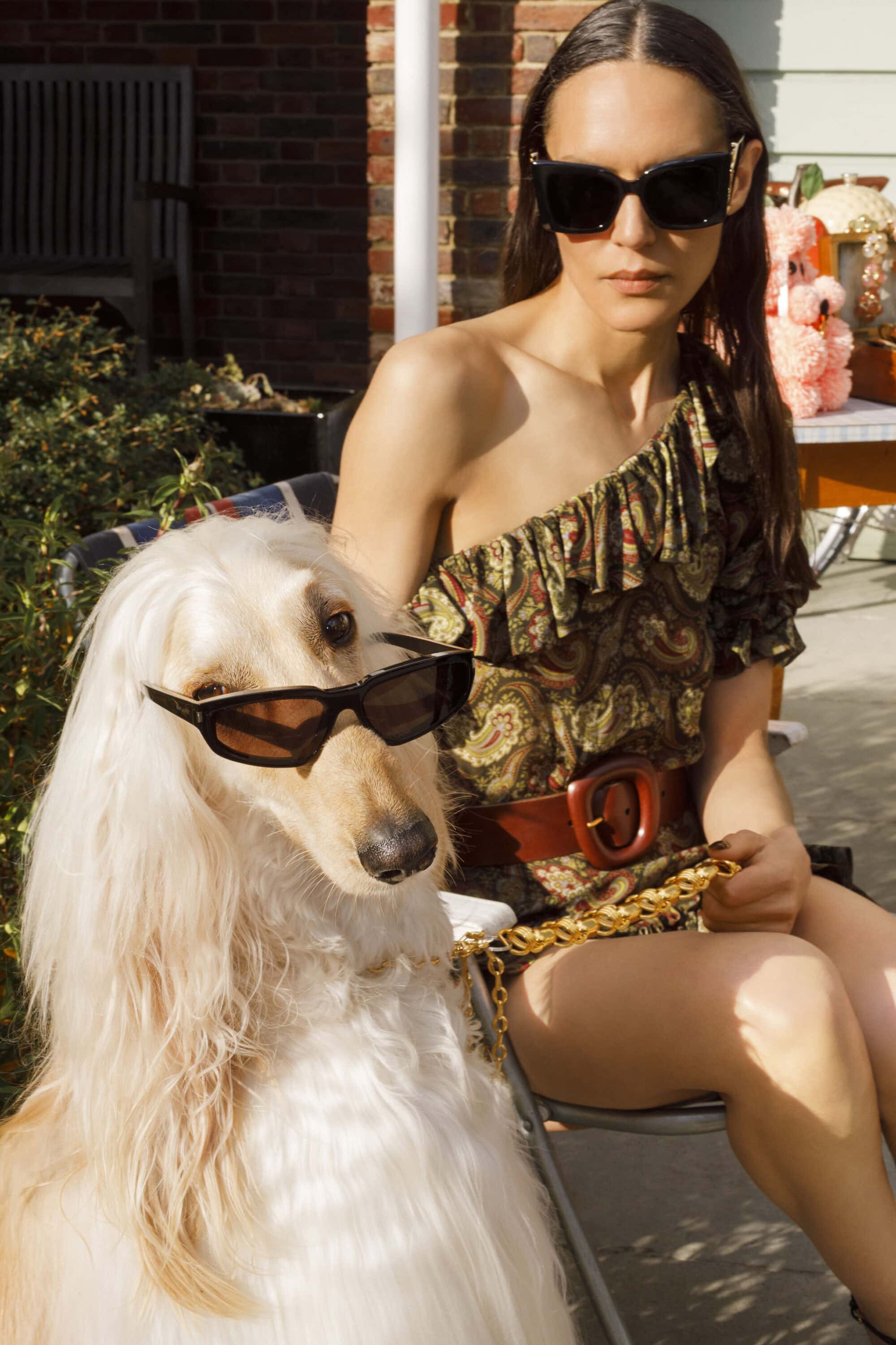 Woman wearing sunglasses and a patterned one-shoulder dress seated next to a long-haired dog wearing sunglasses outdoors.