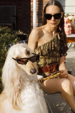 Woman wearing sunglasses and a patterned one-shoulder dress seated next to a long-haired dog wearing sunglasses outdoors.