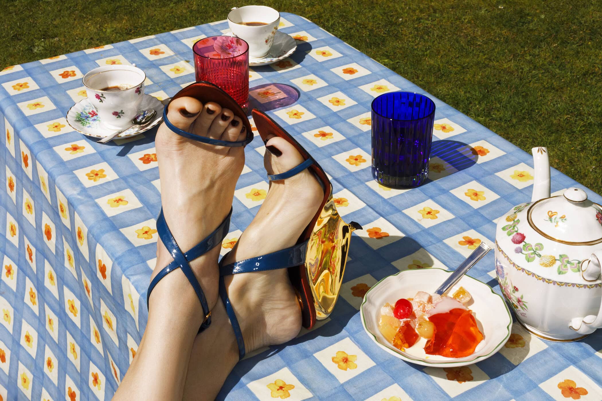Close-up of feet wearing blue strappy sandals propped up on a table with a blue and white checkered tablecloth, teacups, and dessert.