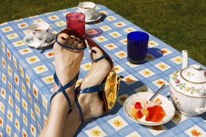 Close-up of feet wearing blue strappy sandals propped up on a table with a blue and white checkered tablecloth, teacups, and dessert.