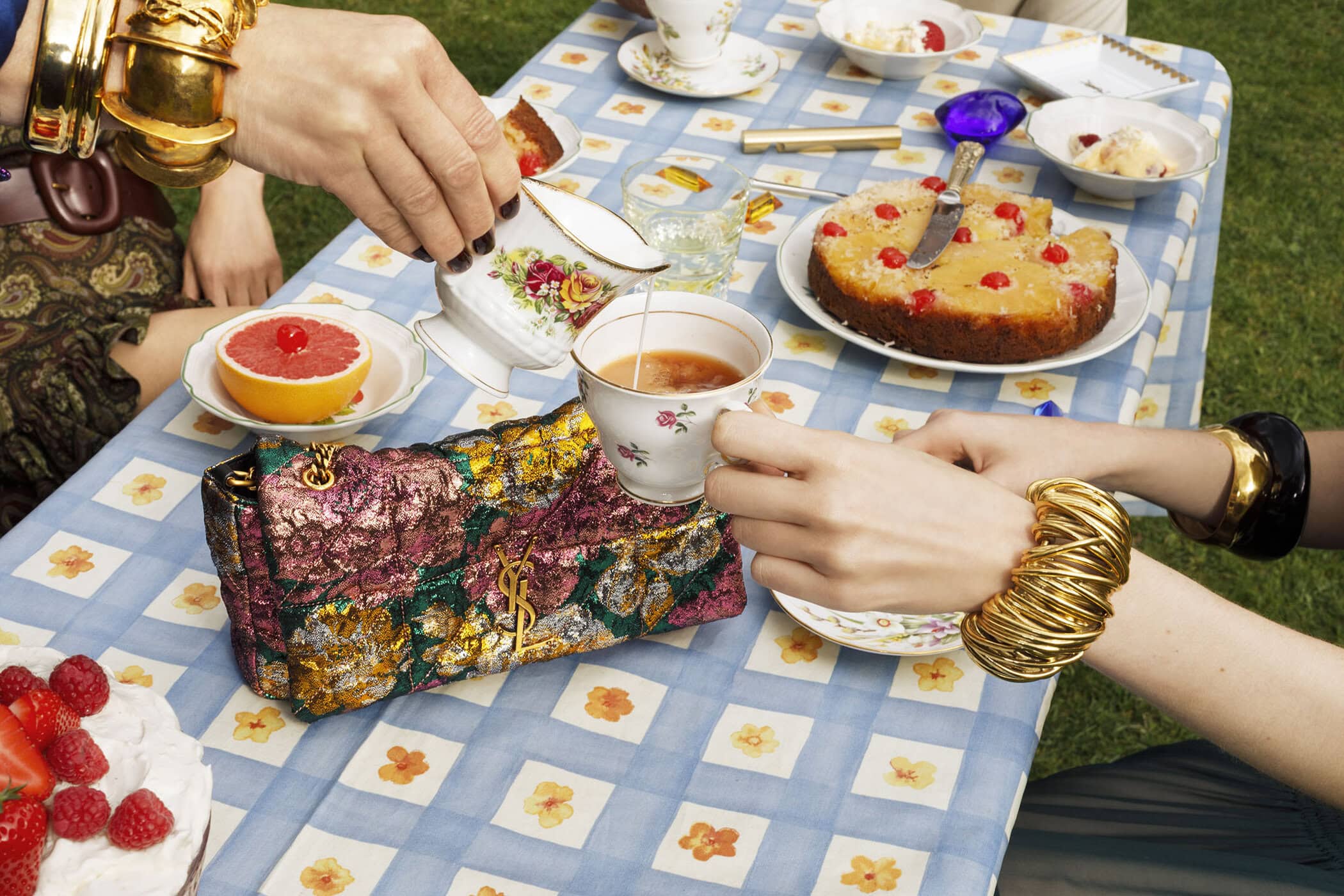 Close-up of hands pouring milk into tea at a picnic table with a patterned cloth, cake, and a Saint Laurent handbag.
