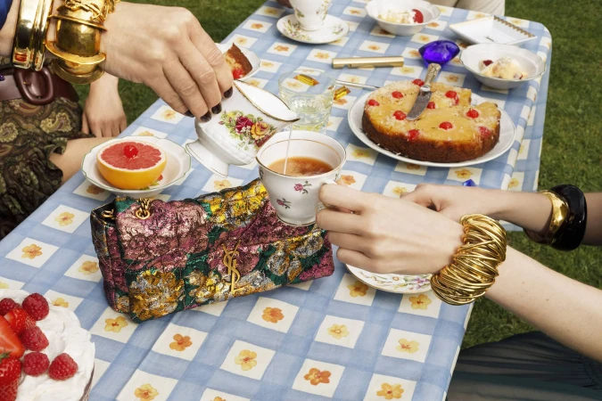 Close-up of hands pouring milk into tea at a picnic table with a patterned cloth, cake, and a Saint Laurent handbag.