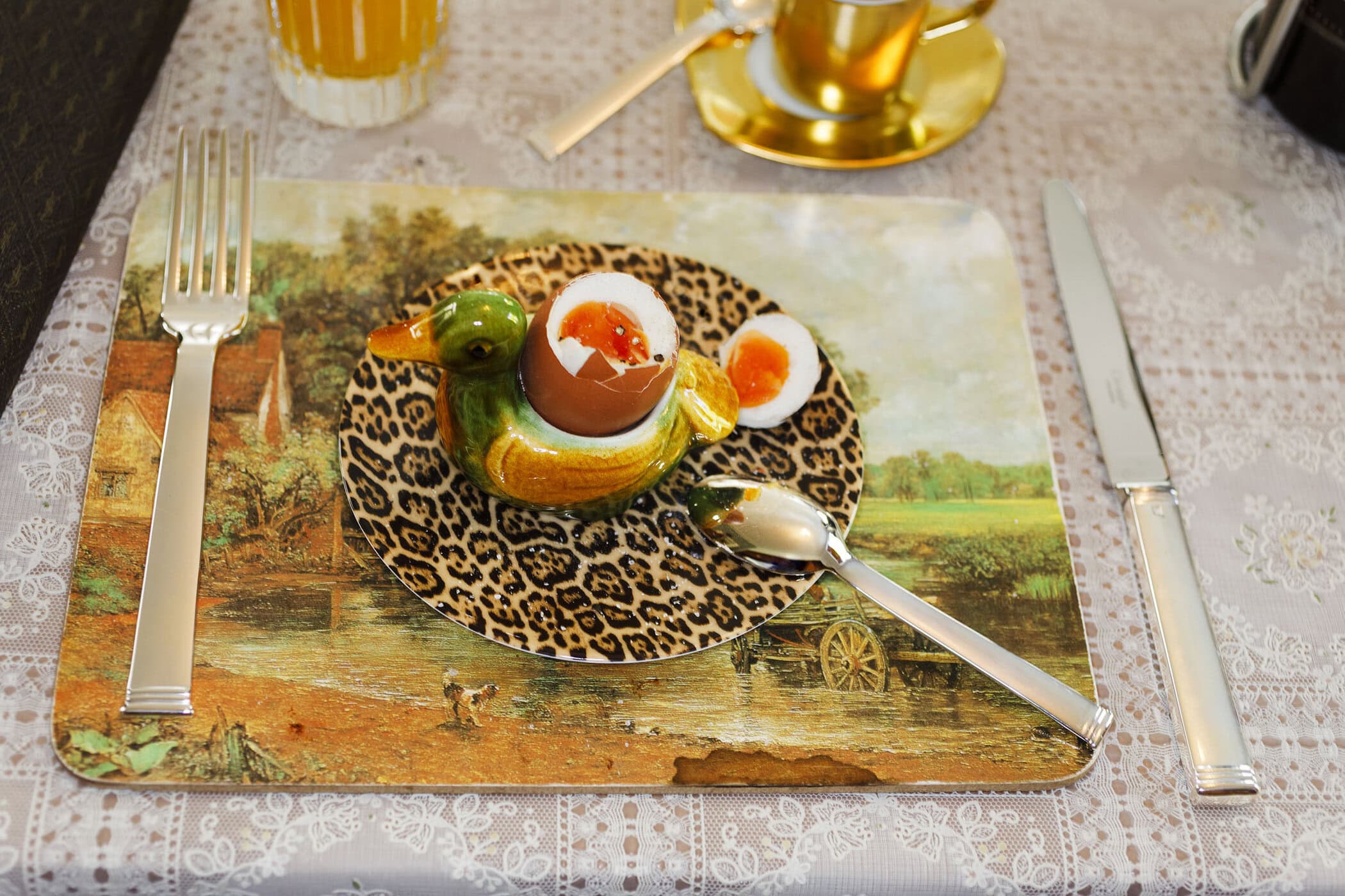 Soft-boiled egg in a duck-shaped egg cup on a patterned plate with a fork and knife on a decorated placemat