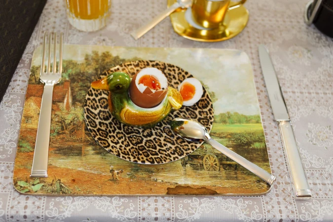 Soft-boiled egg in a duck-shaped egg cup on a patterned plate with a fork and knife on a decorated placemat
