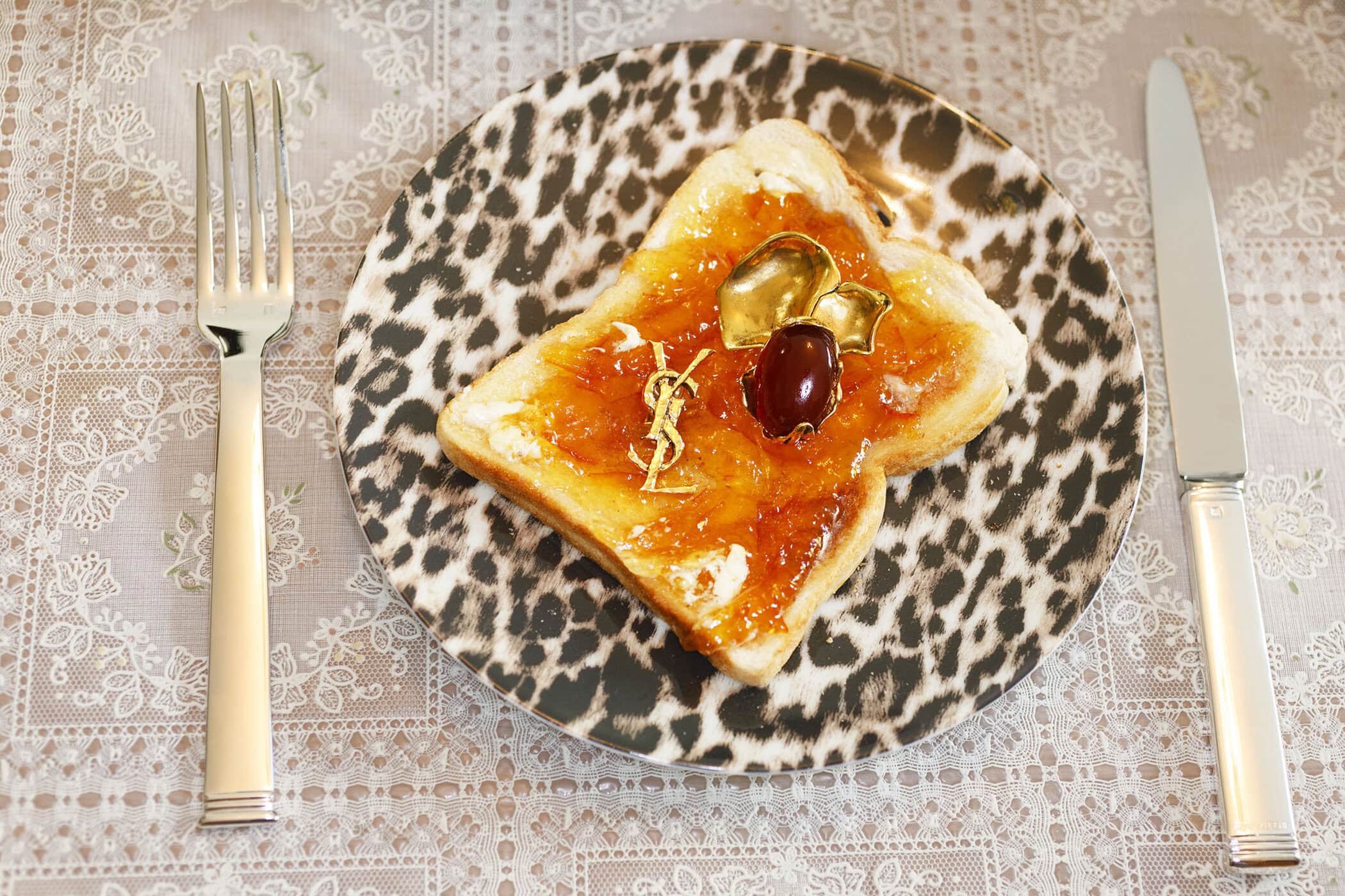 Slice of toast with jam and gold brooch on a leopard print plate flanked by a fork and knife on a lace tablecloth.