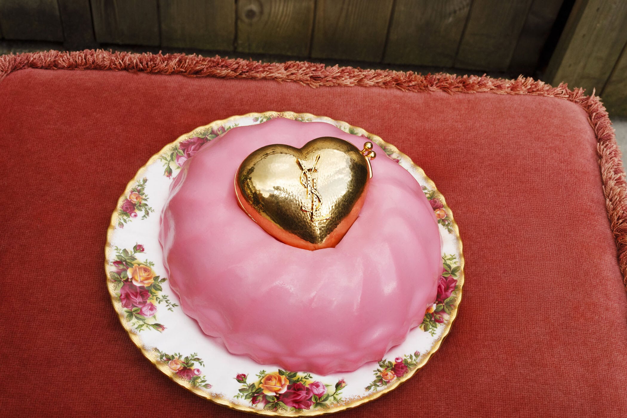 Golden heart-shaped purse atop a pink molded dessert on a floral porcelain plate resting on a textured red surface