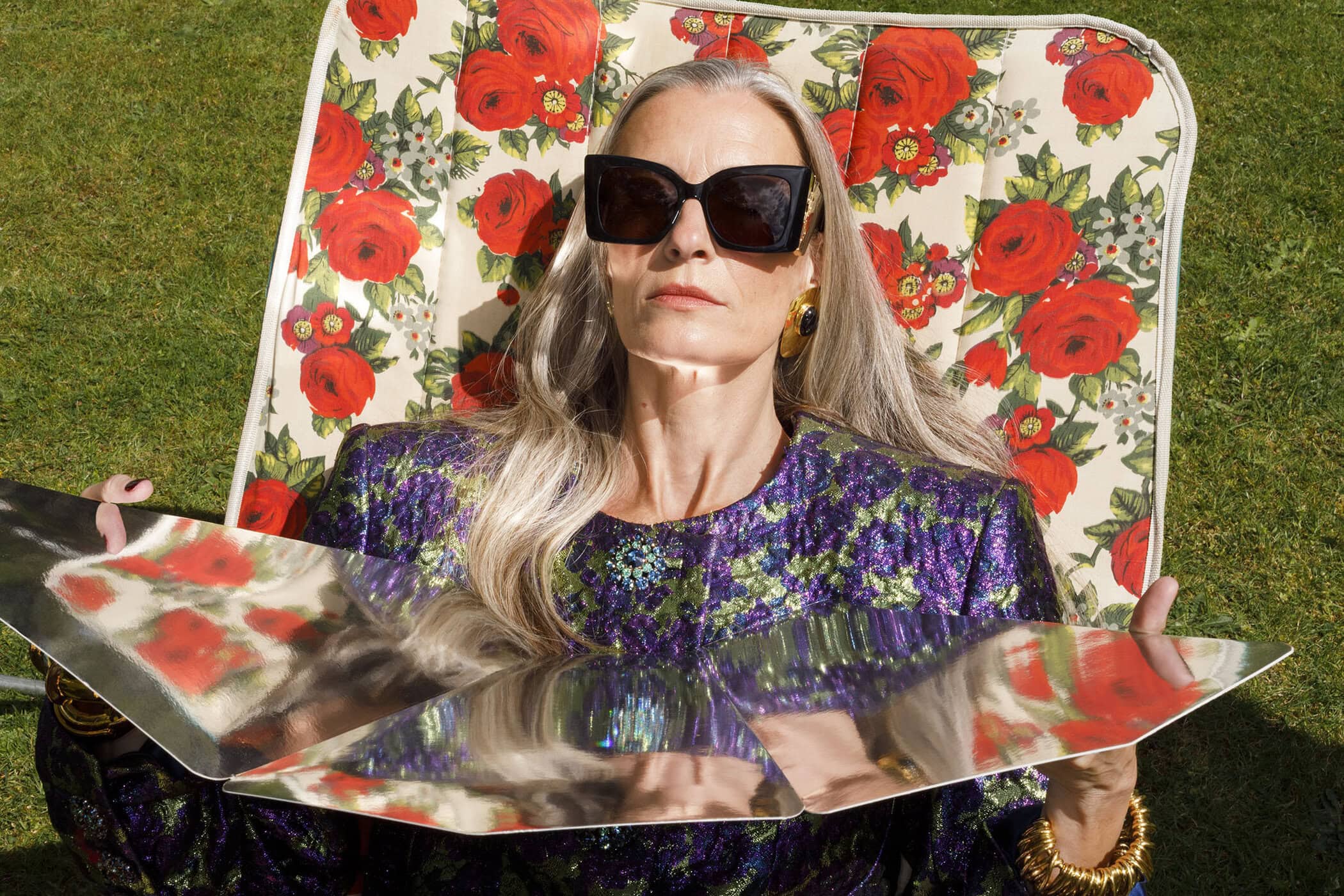 An older woman with long grey hair wearing large black sunglasses and a floral patterned top relaxing in a floral lounge chair outdoors.