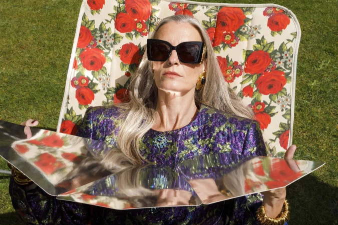 An older woman with long grey hair wearing large black sunglasses and a floral patterned top relaxing in a floral lounge chair outdoors.