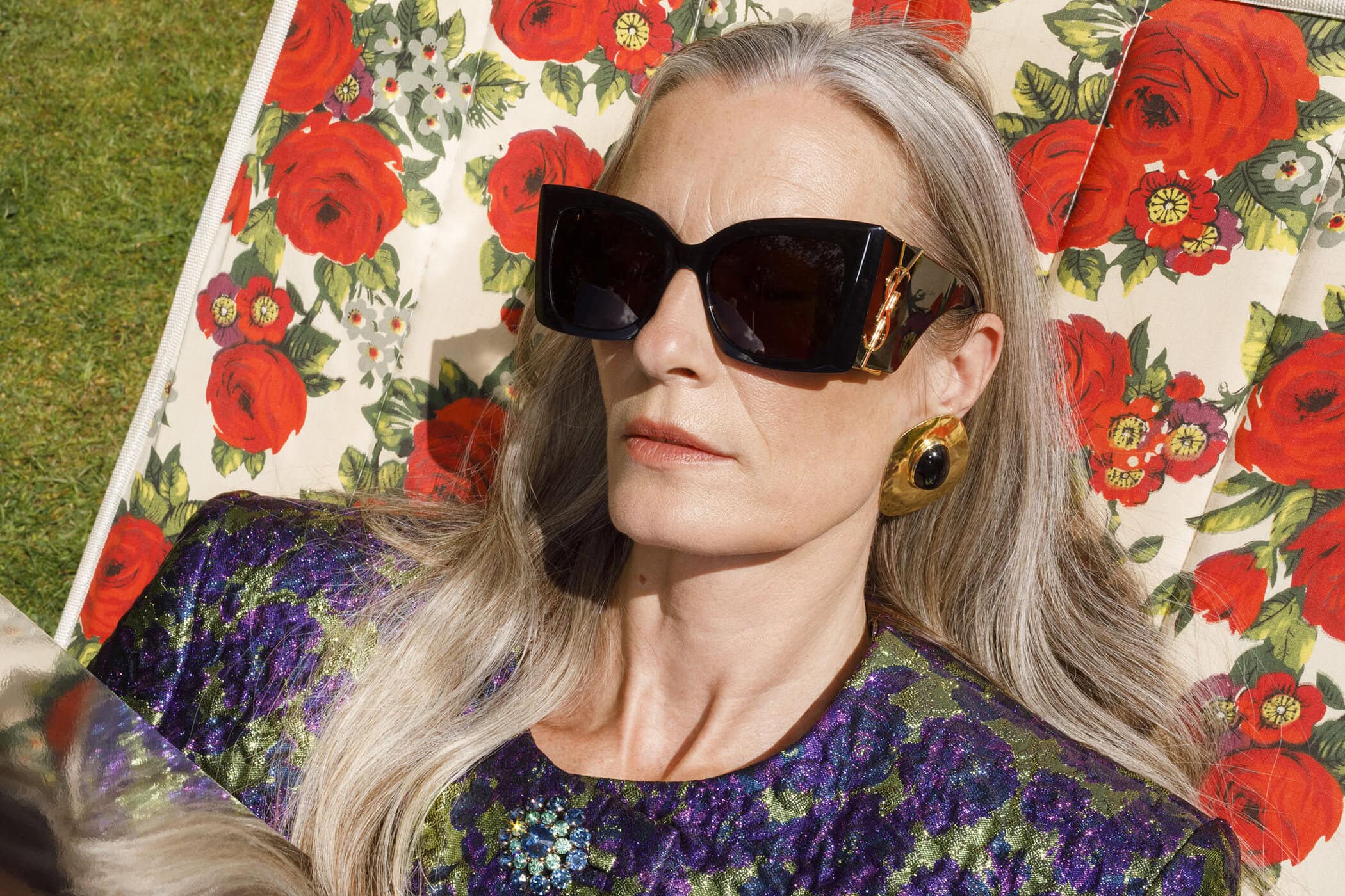 An elderly woman with long grey hair wearing black sunglasses and floral pattern top reclining on a matching floral deckchair.