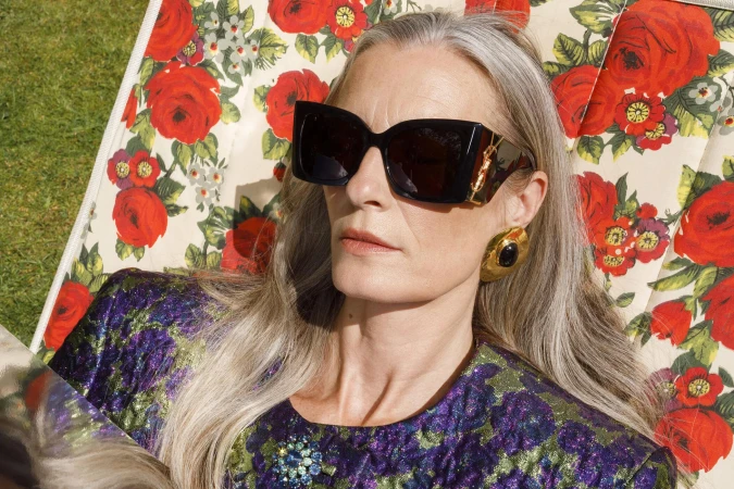 An elderly woman with long grey hair wearing black sunglasses and floral pattern top reclining on a matching floral deckchair.