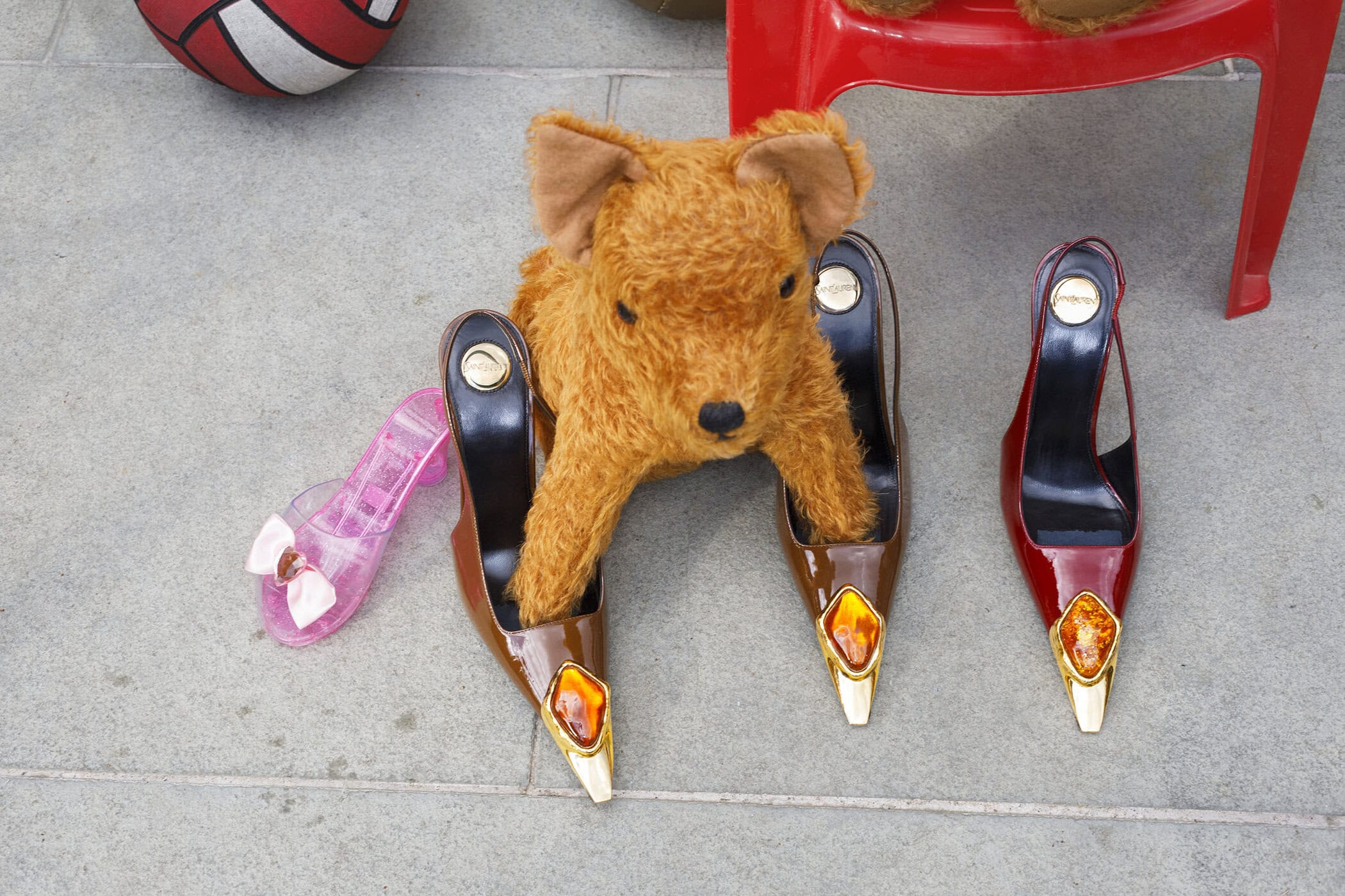 Fluffy brown toy dog standing inside two brown patent leather pumps with gold gem toes on grey stone pavement.