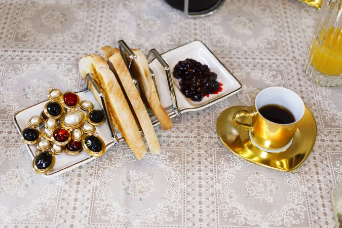 Toast rack with sliced bread, jam, gold coffee cup, and jewelry displayed on a patterned tablecloth.