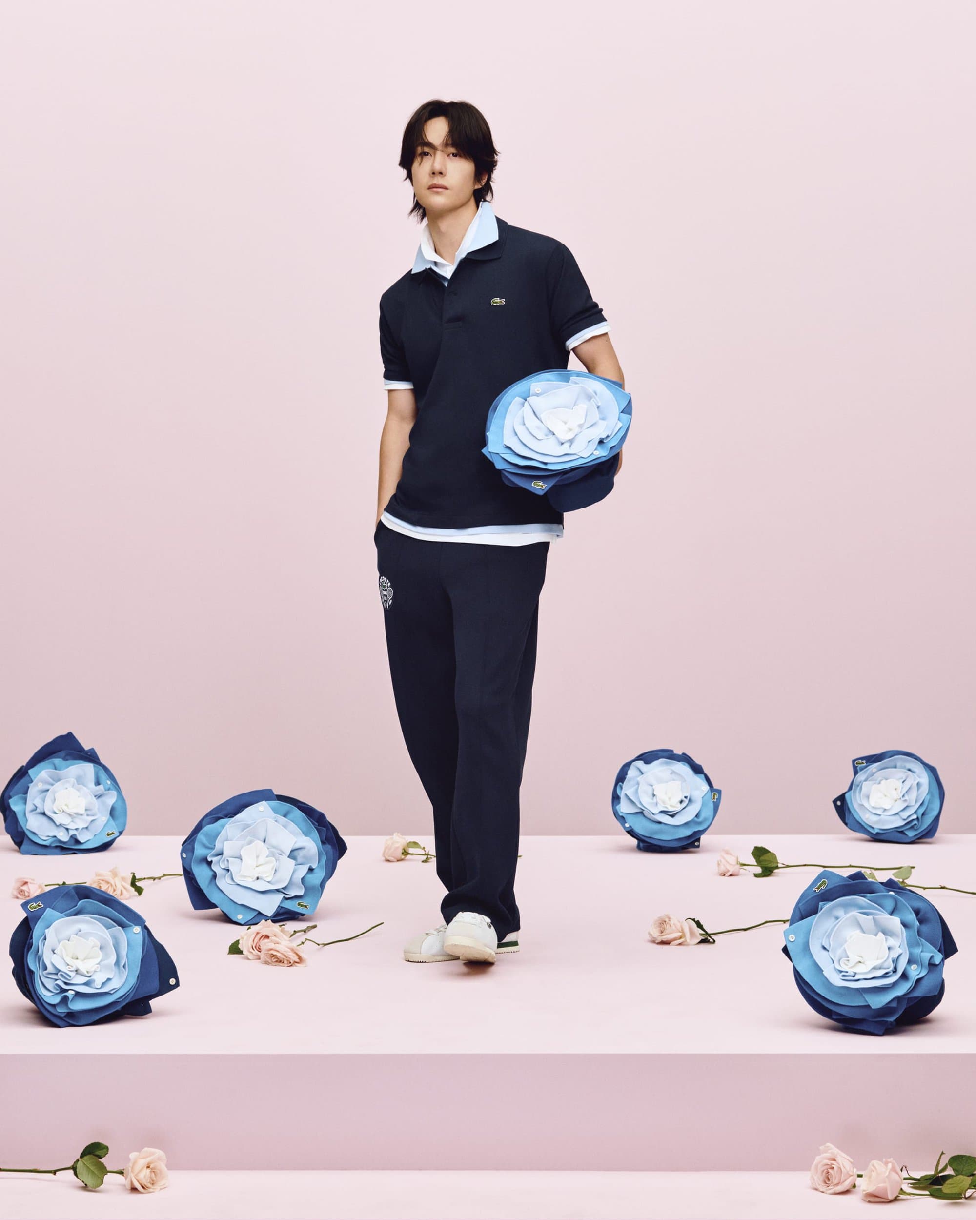 Wang Yibo in navy polo shirt and matching trousers holding large blue paper flower against pink background