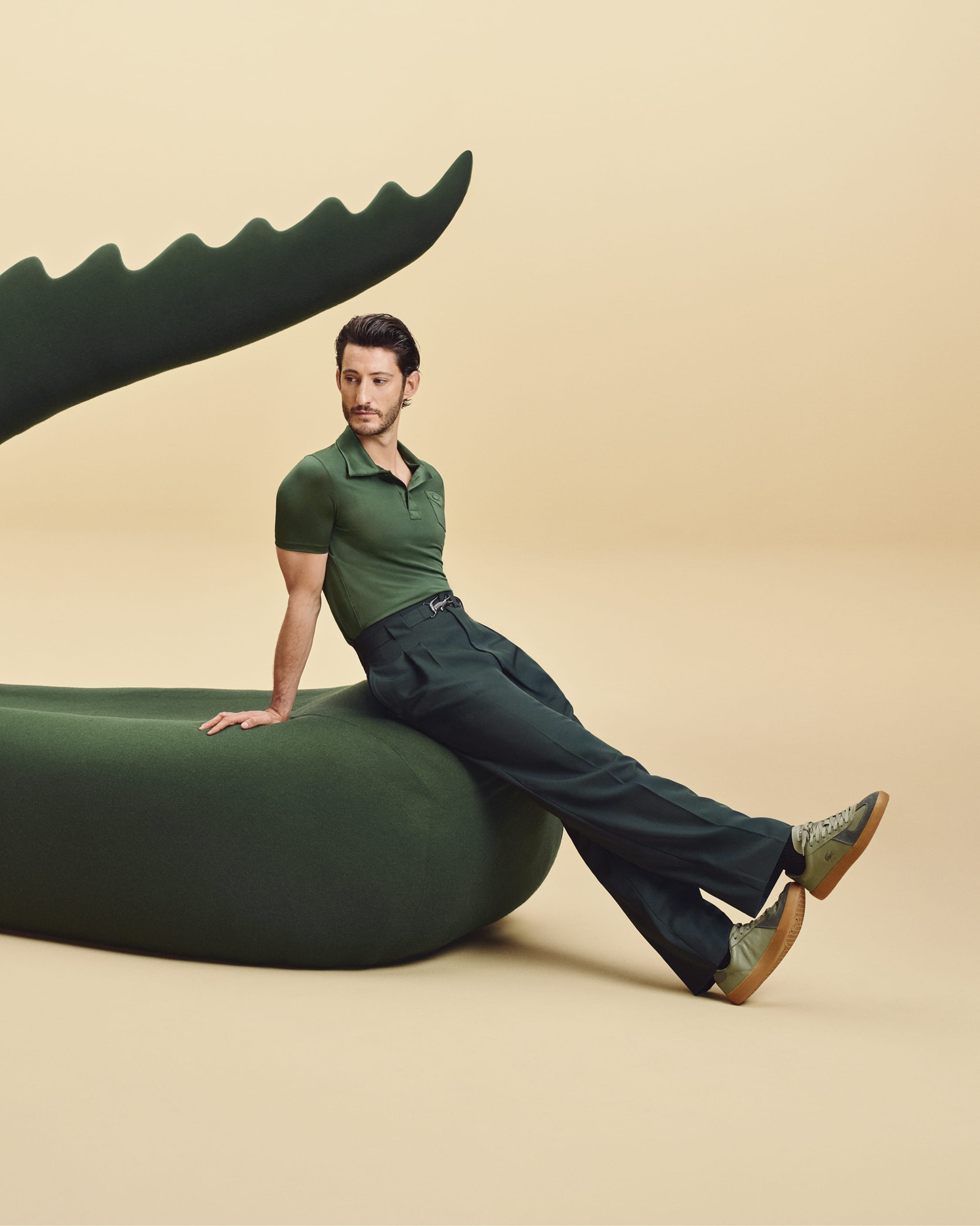 Man seated on a large dark green sculptural prop against a beige background for Lacoste Play with Icons campaign