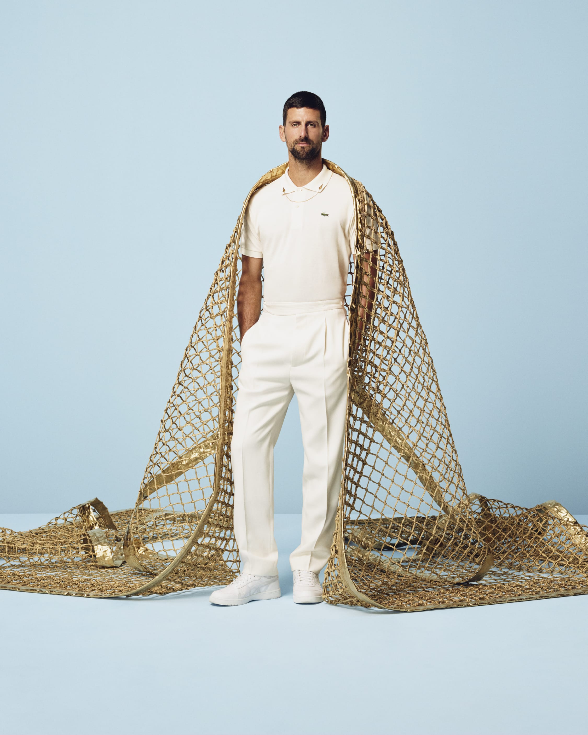 Novak Djokovic wearing white polo and trousers wrapped in a large gold mesh net on blue background