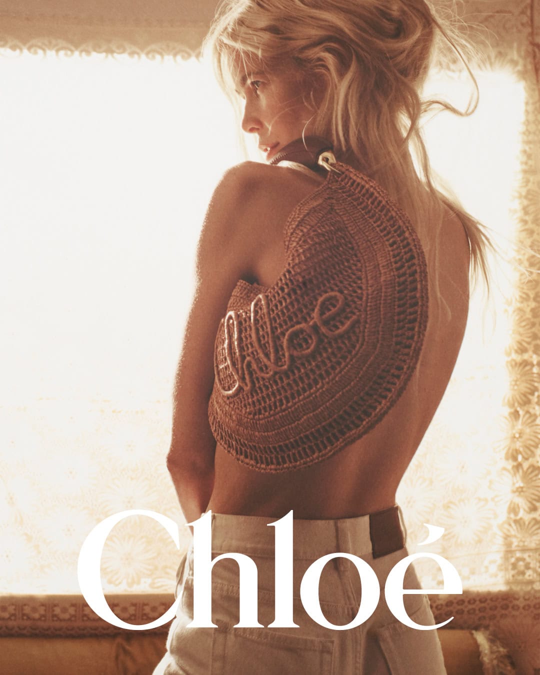 Claudia Schiffer posing for Chloé High Summer 2025 ad campaign, wearing a woven shoulder bag against a bright window backdrop.