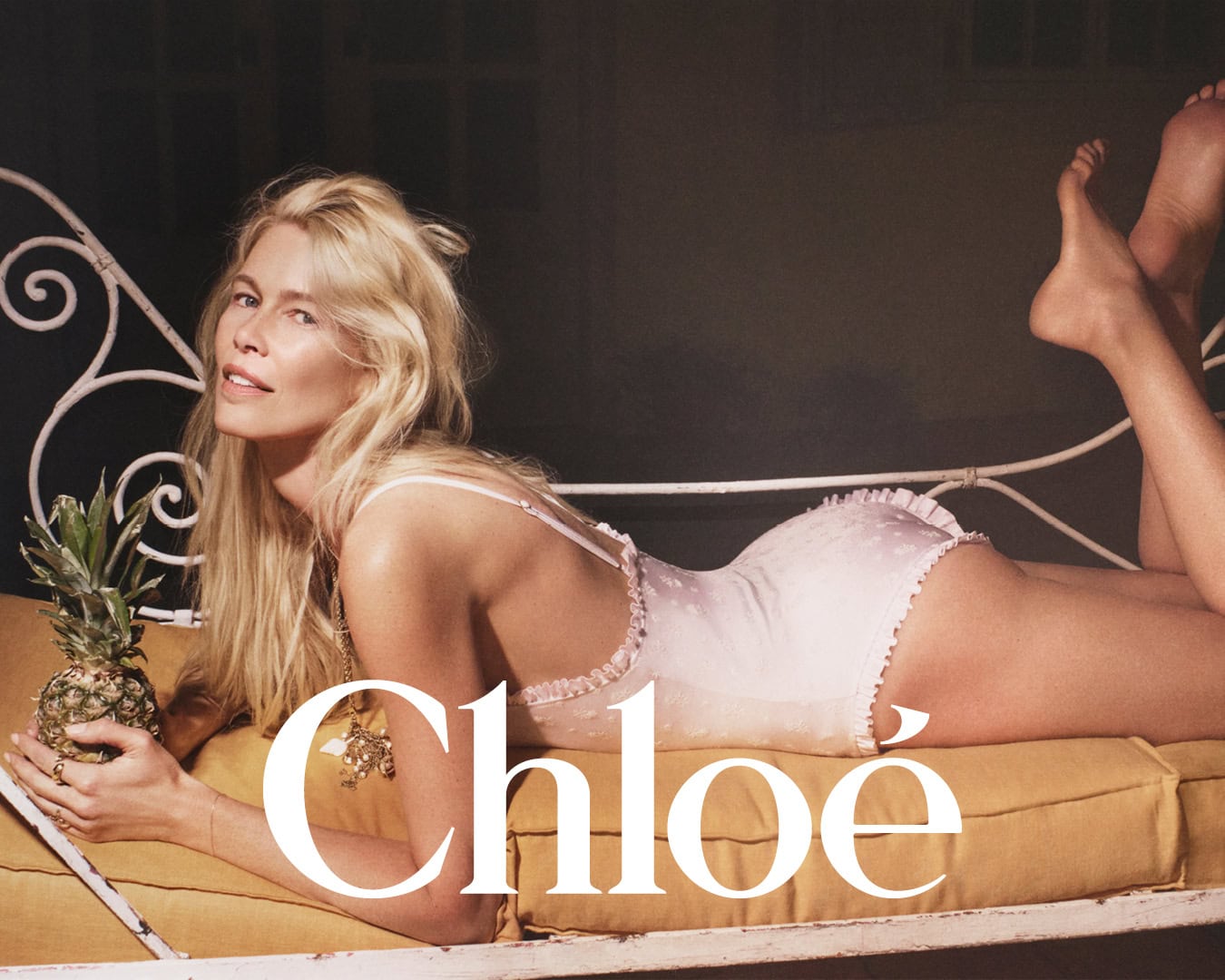 Claudia Schiffer lying on a bed holding a small pineapple wearing a light pink swimsuit for Chloe High Summer campaign