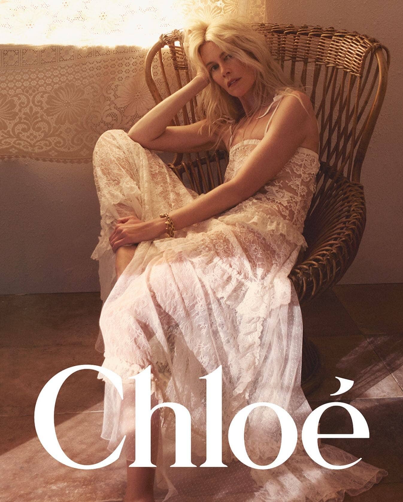Claudia Schiffer seated in a wicker armchair wearing a white lace dress for the Chloe High Summer 2025 ad campaign.