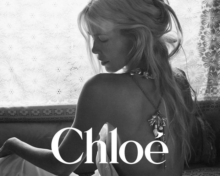 Claudia Schiffer in a black and white campaign image wearing back jewelry against a patterned curtain.