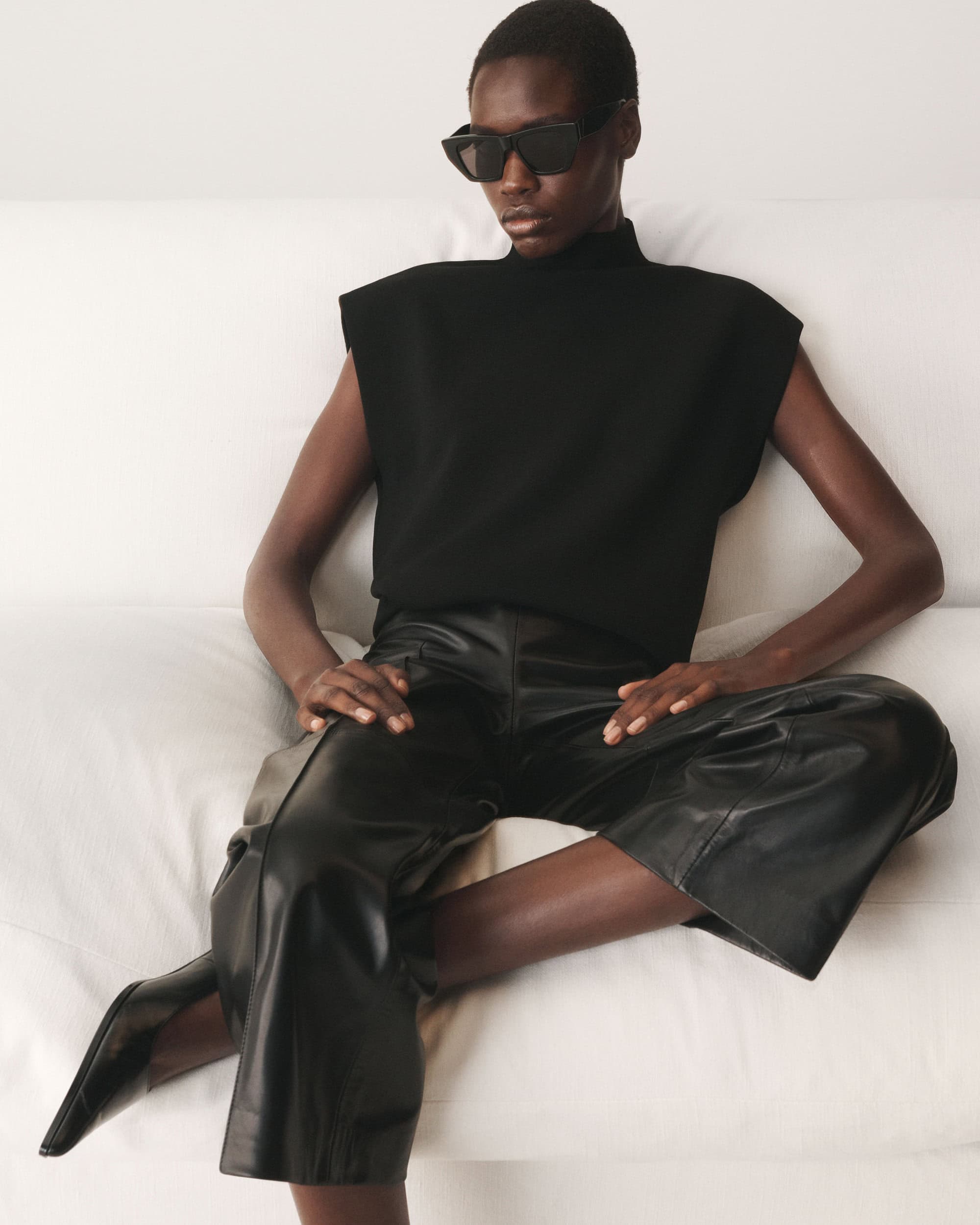 Black woman seated wearing a black sleeveless top, leather trousers, and dark sunglasses against a white background
