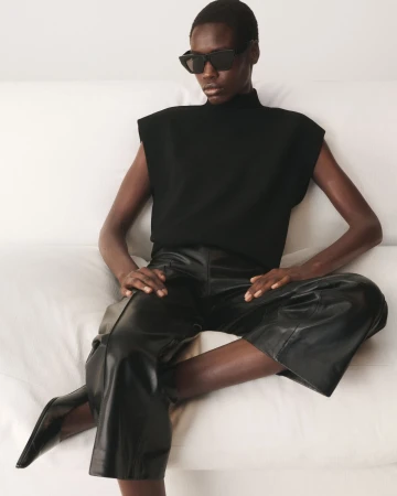 Black woman seated wearing a black sleeveless top, leather trousers, and dark sunglasses against a white background