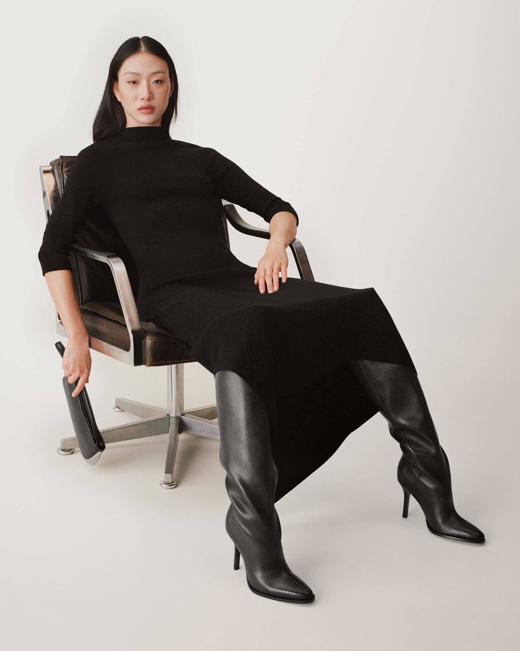 Asian woman seated in a leather armchair wearing a black dress and black leather boots against a white studio background.