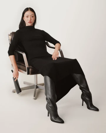 Asian woman seated in a leather armchair wearing a black dress and black leather boots against a white studio background.